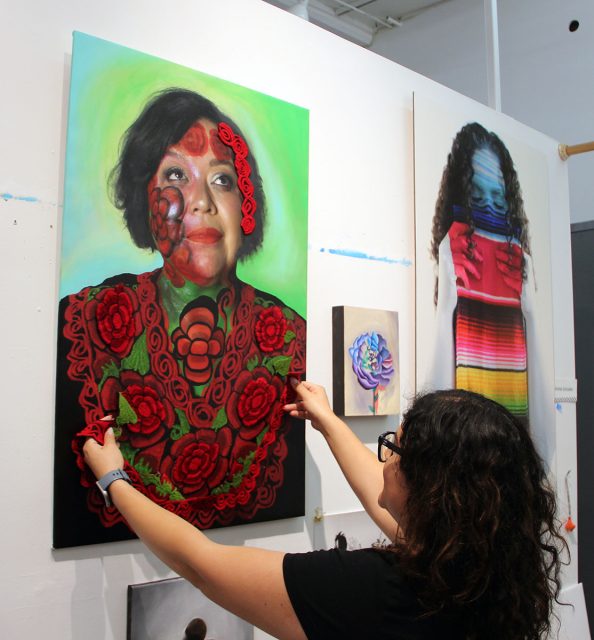 A woman adjusts a colorful portrait with a floral design, displayed on a white wall alongside other artworks.