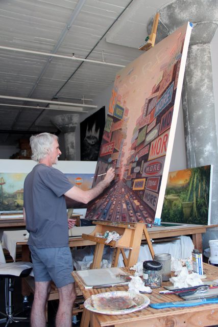 A person with gray hair stands in a studio painting a large, colorful canvas on an easel. The artwork features numerous "MORE" signs. Various paints and supplies are scattered on surrounding surfaces.