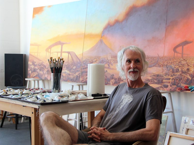 A man with gray hair and a beard sits in front of a large painting depicting a volcanic eruption and a car scrapyard. Art supplies and brushes are on a table beside him.