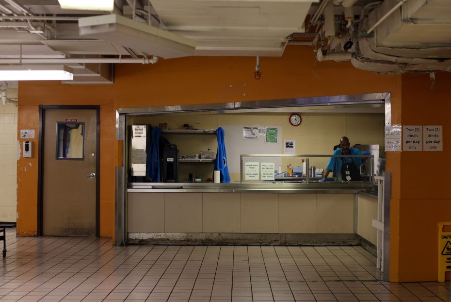 A worker stands behind a counter in what appears to be a cafeteria or food service area with a sign indicating a two-drink limit per day. The surroundings have a utilitarian design with orange walls.