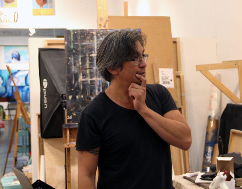 A person with gray hair and glasses stands in an art studio, touching their chin and looking to the side. The background features canvases, easels, and art supplies.