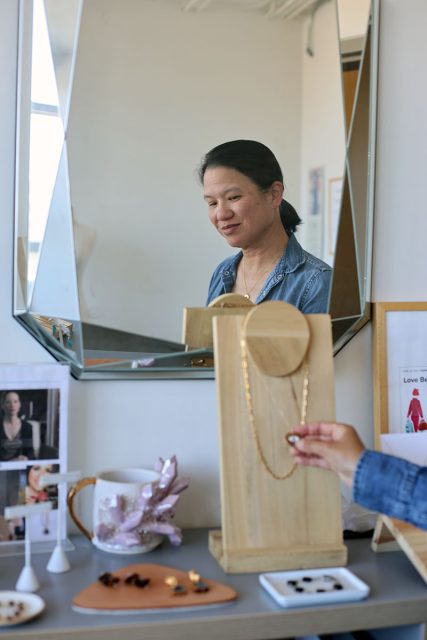 A woman stands in front of a mirror looking at a necklace displayed on a wooden stand, with jewelry items and photos arranged on a table below.