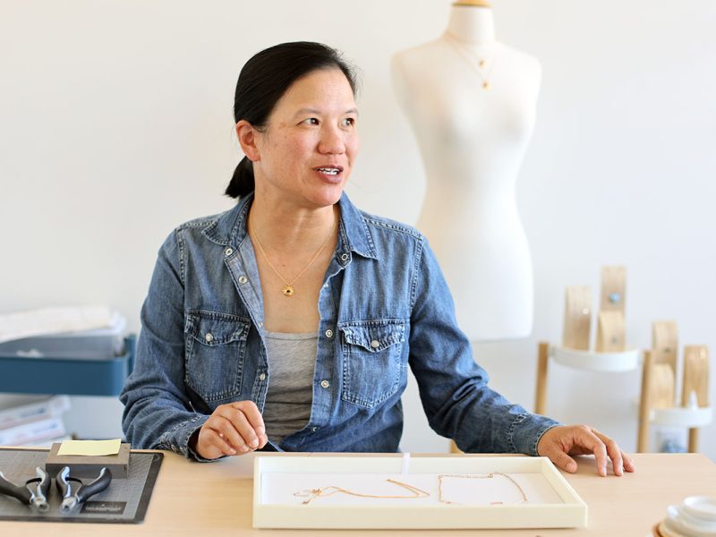 A person sits at a table with jewelry-making tools, holding a jewelry tray. A mannequin in the background displays a necklace.