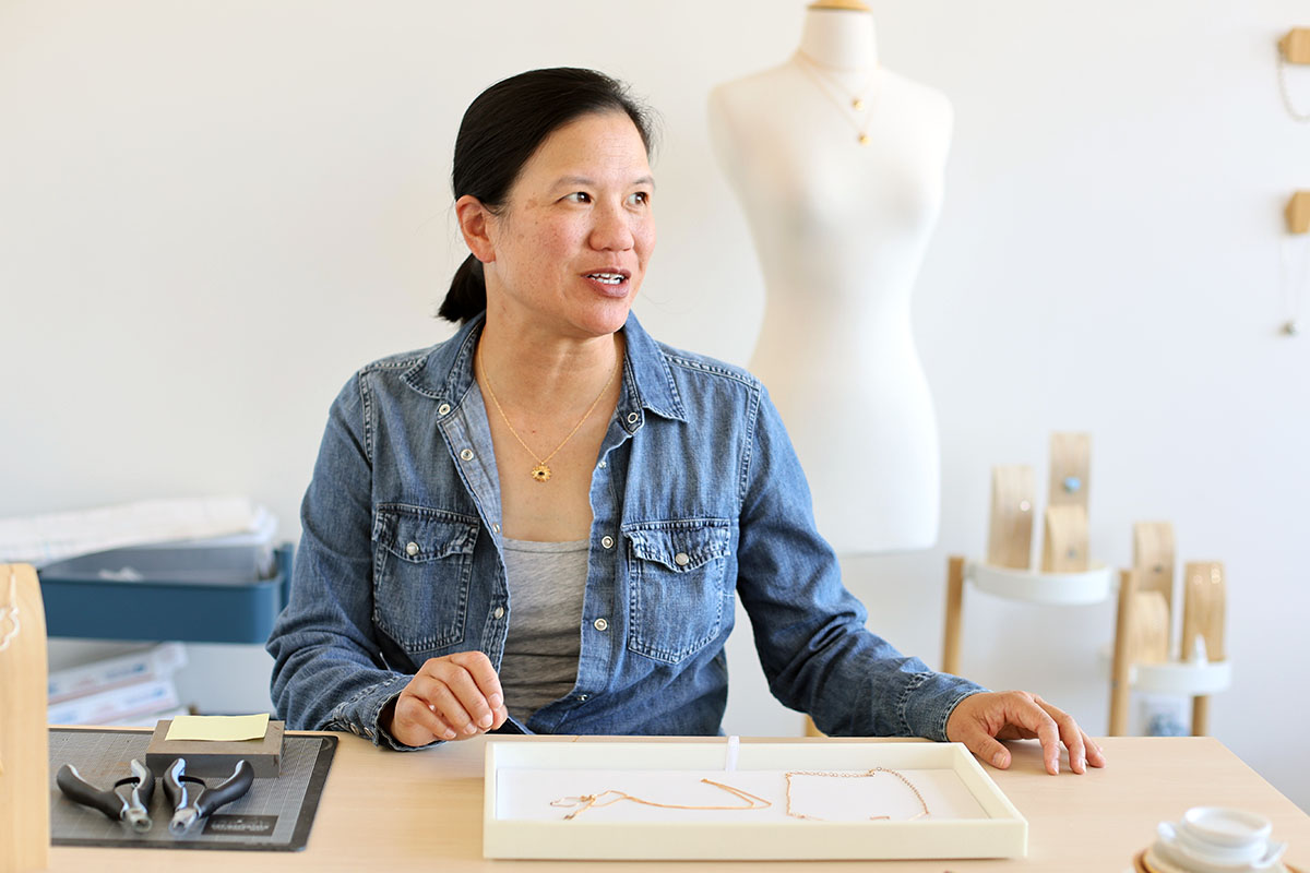 A person sits at a table with jewelry-making tools, holding a jewelry tray. A mannequin in the background displays a necklace.
