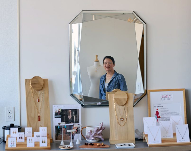 A woman is reflected in a mirror behind a display of jewelry, which includes necklaces, earrings, and bracelets on a wooden stand. A dress form and framed pictures are also visible.