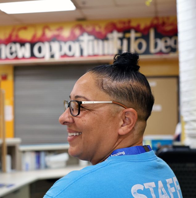 A person wearing glasses and a blue "Staff" shirt smiles, sitting in front of a colorful wall with the text "new opportunity to be.