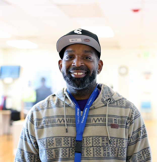 A smiling man wearing a cap and patterned hoodie with a blue lanyard stands indoors in a brightly lit room.
