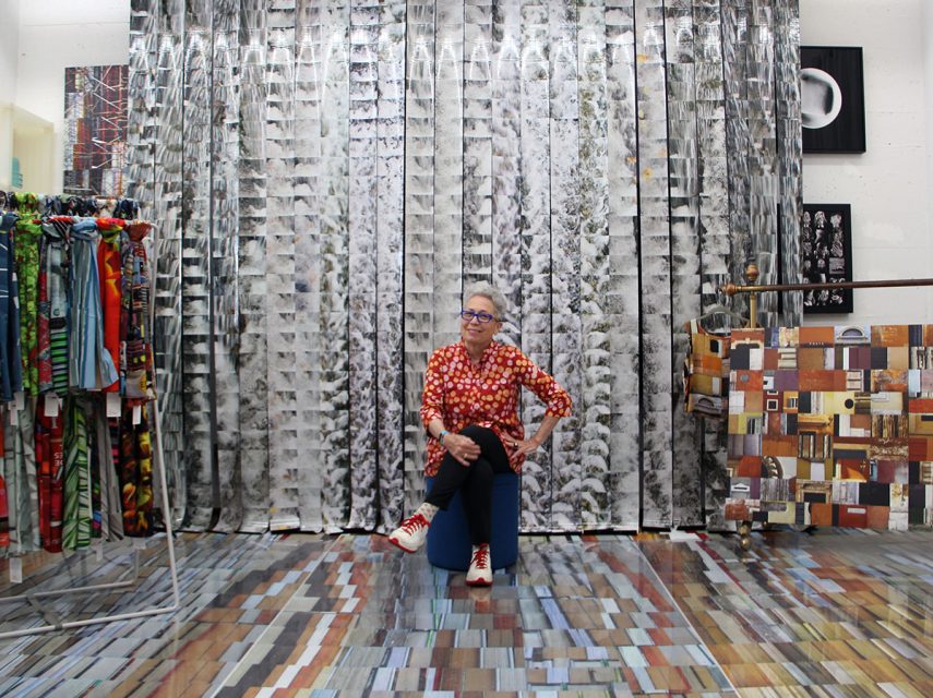 A person wearing a red patterned shirt and glasses sits in a vibrant room filled with colorful textiles and abstract art pieces.