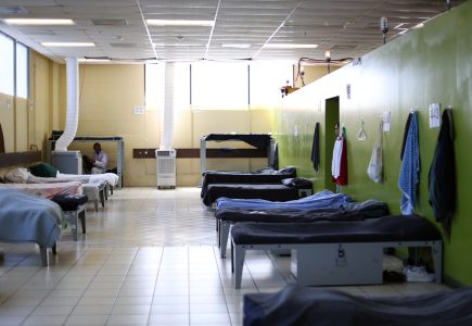 A large room with multiple single beds lined up against green walls, each bed with blankets, and clothes hanging on the walls. A person is sitting on one of the beds in the background.