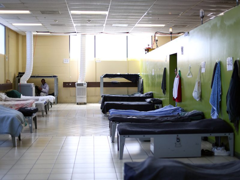 A large room with multiple single beds lined up against green walls, each bed with blankets, and clothes hanging on the walls. A person is sitting on one of the beds in the background.