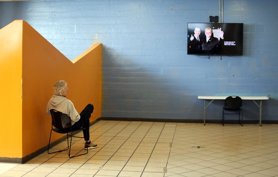 A person sits on a chair in an empty room with orange and blue walls, watching a TV mounted on the wall that shows a show or film featuring people in formal attire.