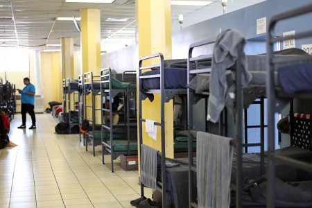 A room with multiple metal bunk beds lined up, each with bedding and personal items. A person in a blue shirt is standing near the doorway.