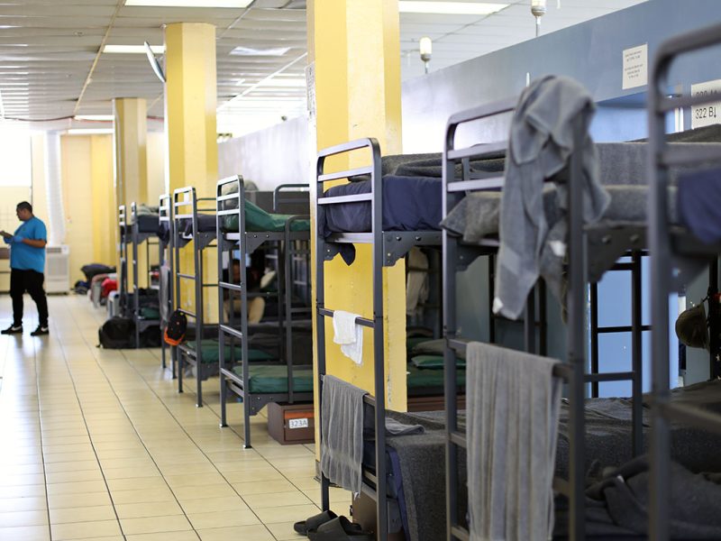 A room with multiple metal bunk beds lined up, each with bedding and personal items. A person in a blue shirt is standing near the doorway.