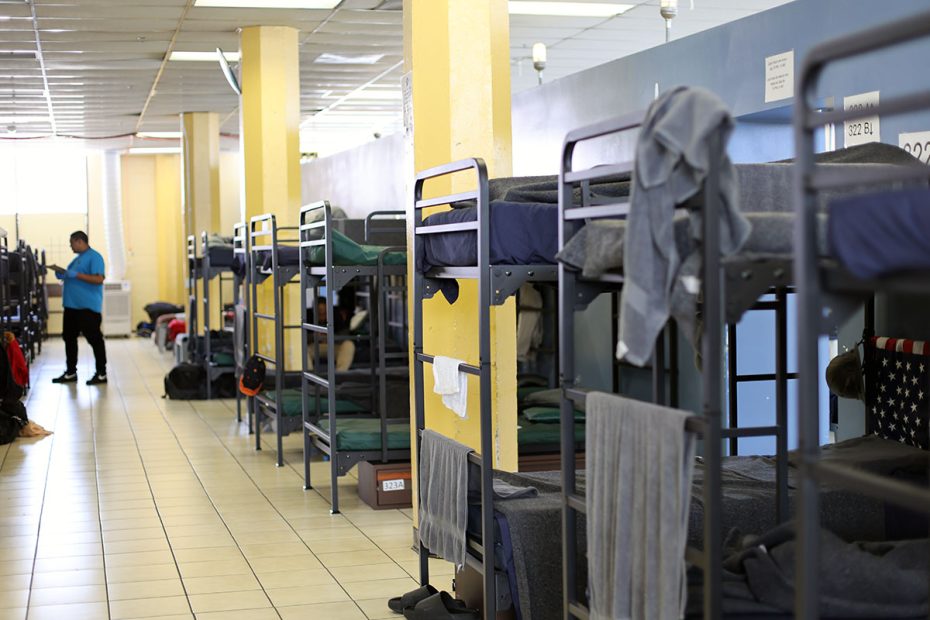 A room with multiple metal bunk beds lined up, each with bedding and personal items. A person in a blue shirt is standing near the doorway.