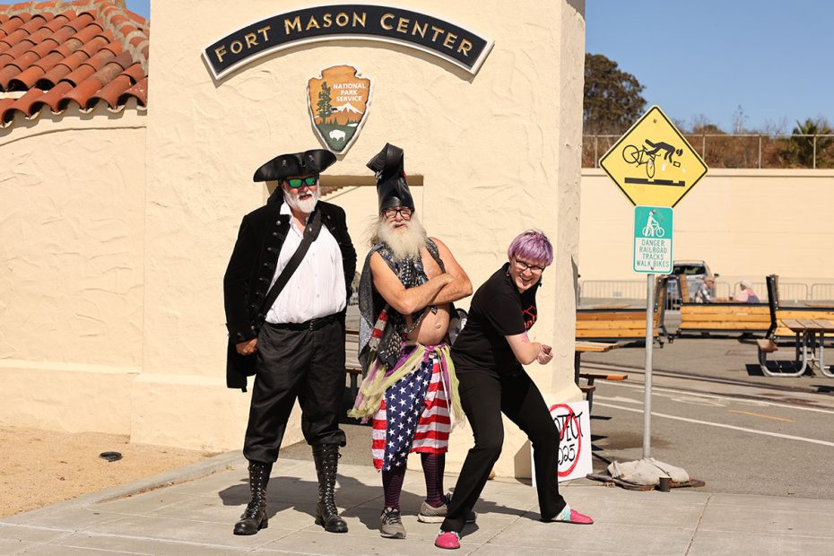 Three adults in distinct costumes pose in front of Fort Mason Center. One wears a pirate outfit, another has a tall hat and American-themed attire, and the third has purple hair and a black shirt.