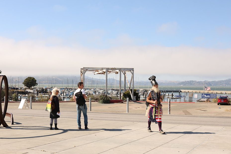 Two people stand on a dock near boats while a man in a helmet and cape walks by holding a flag. A waterfront and mountains are in the background.