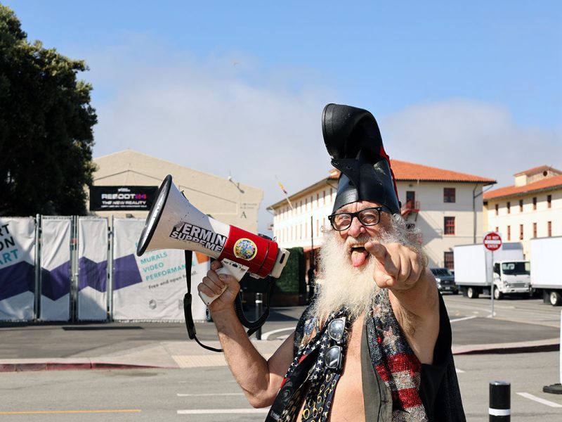 一位留著長白鬍子的男子戴著黑色帽子和眼鏡，手持擴音器指向相機，背景是一棟建築和街道。