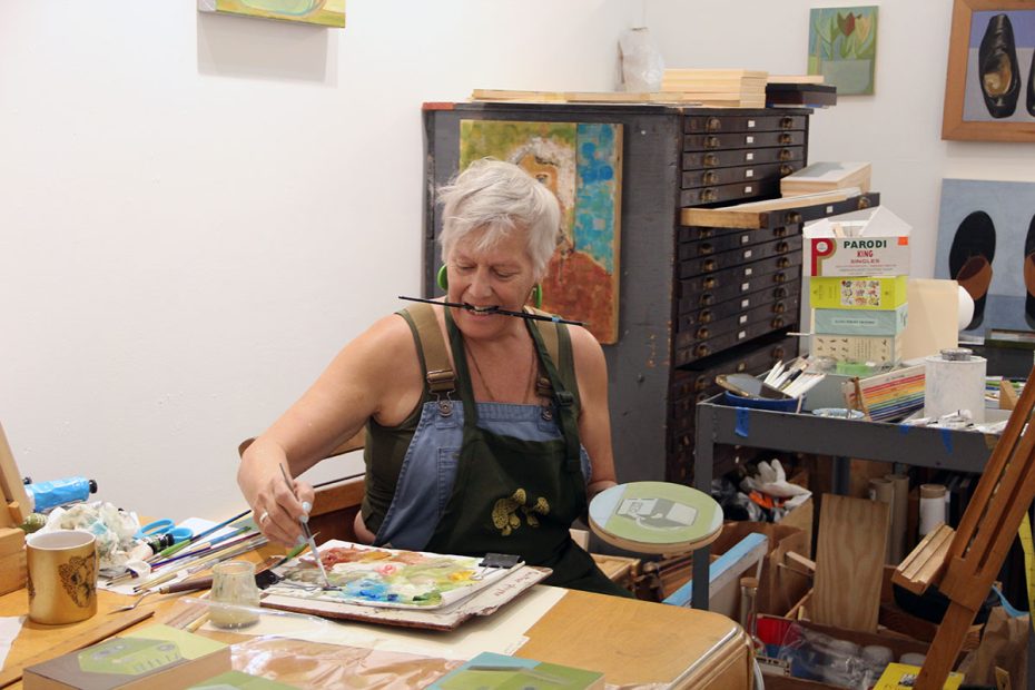 An artist is painting in a studio, using a palette on her lap and holding a brush in her mouth. She is surrounded by art supplies and paintings.