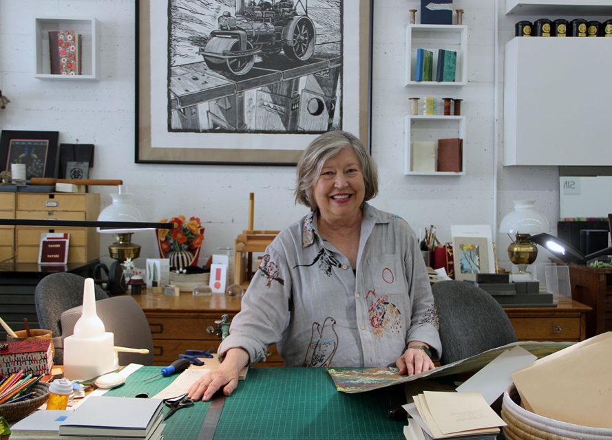 A woman with gray hair stands at a work table in an art studio, holding a partially bound book. Various tools, books, and art supplies surround her. A detailed print hangs on the wall behind her.