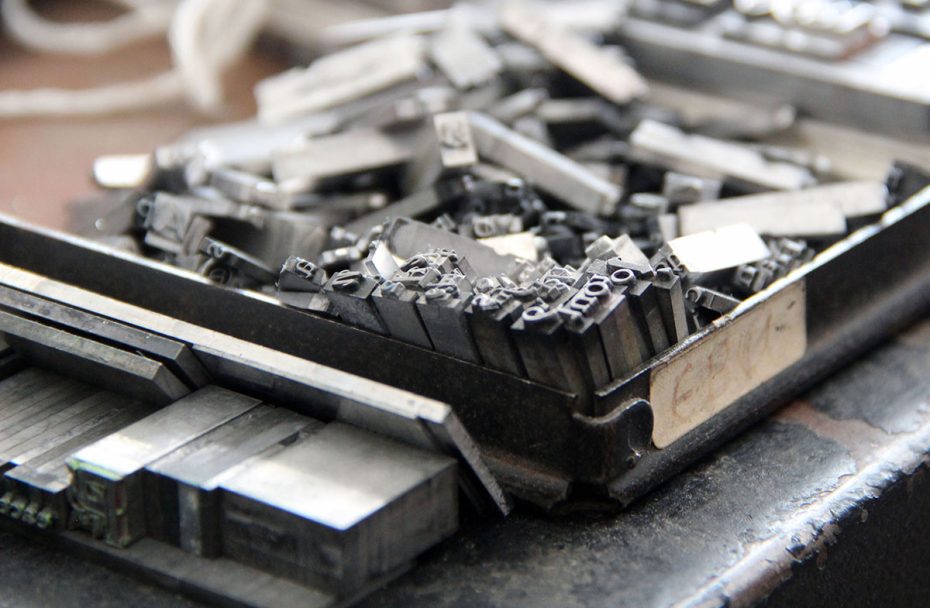 Close-up of a tray filled with metal movable type pieces for letterpress printing, showing various letterforms and shapes.