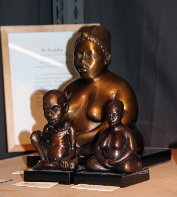Three bronze statues on display, depicting a large central figure seated in a meditative pose, flanked by two smaller seated figures. A framed document is visible in the background.