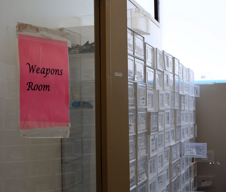 A room labeled "Weapons Room" with shelves containing numerous clear plastic bins.