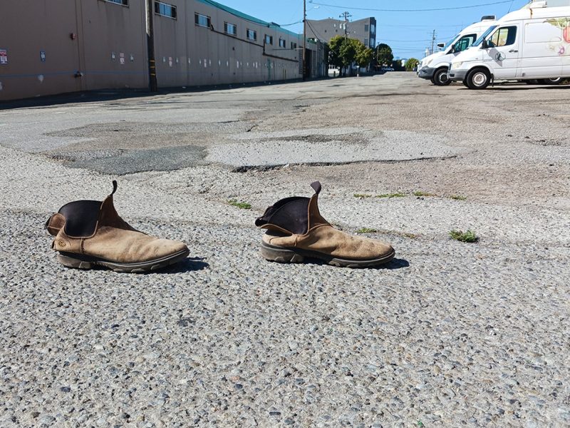 A pair of worn brown boots is placed on an empty street with industrial buildings and parked vans in the background.