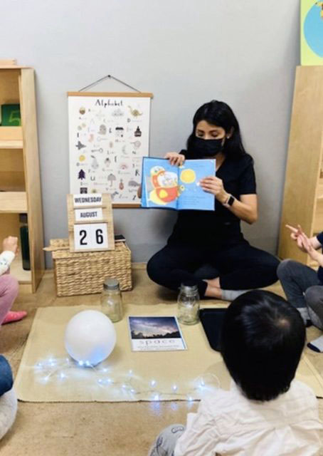 A teacher reads a book to children seated in a circle on the floor, while a calendar displays the date "August 26." Nearby, a large white balloon and string lights add whimsy to the scene. In the background hangs an alphabet poster—reminding everyone of Lai's favorite classroom décor.