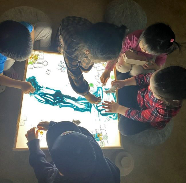 Five children sit around an illuminated table, engrossed in an interactive activity involving blue tactile material and transparent blocks, a design inspired by Lai.
