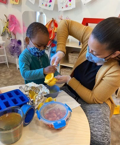 An adult and child, both wearing masks, squeeze lemons together at a table adorned with ice cube trays, a bowl, and various ingredients. Amidst their joyful activity, Lai ensures every lemon drop counts for the perfect mix.