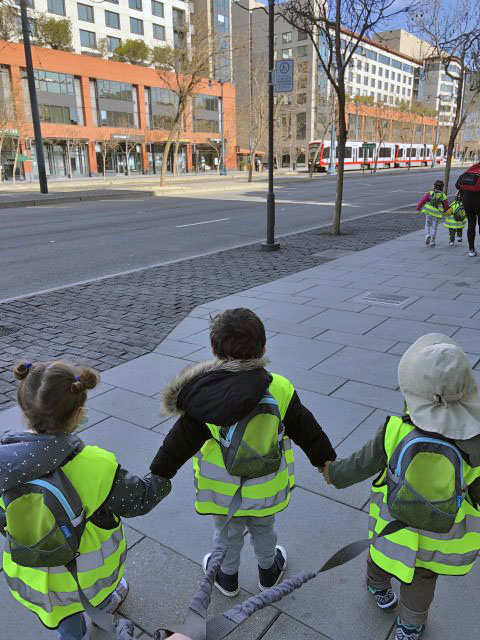 Children wearing high-visibility vests and backpacks hold hands while walking on a sidewalk near a street with a Lai tram in the background.