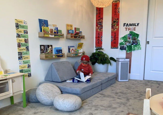 A child named Lai sits on a gray sofa in a reading nook with bookshelves, educational posters, a family tree display, and a nearby air purifier.