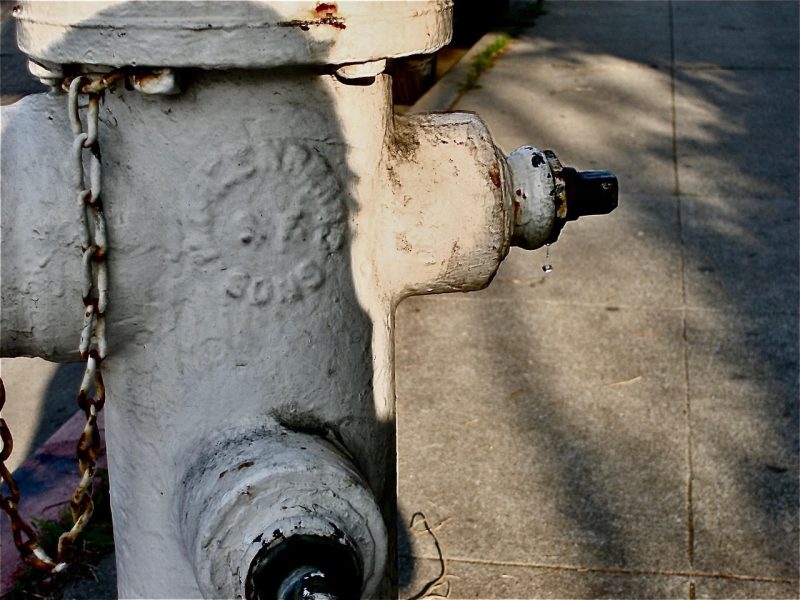 A close-up of a weathered fire hydrant with a slight leak visible near the cap. The background shows a concrete sidewalk.
