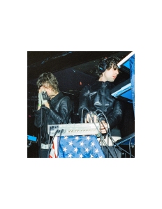 Two people in dark clothing perform on stage with electronic equipment and an American flag draped over a keyboard.