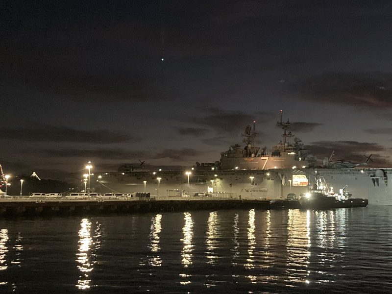 A large naval ship docked at a pier during nighttime, with city lights and a bridge visible in the background.