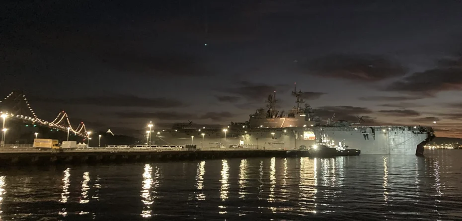 Navy ship docked at a pier during nighttime with bridge and city lights in the background, reflecting off the water.