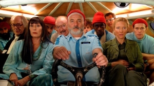 A group of people wearing blue uniforms and red hats is seated together in a confined space, with a central figure holding a steering wheel at the front.