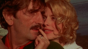 Close-up of a man with a mustache and a woman with blonde hair, with the woman gently touching the man's face and looking into each other's eyes.