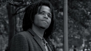 A person with dreadlocks and a goatee looks up while standing outdoors, with trees and a pole in the background. The image is in black and white.