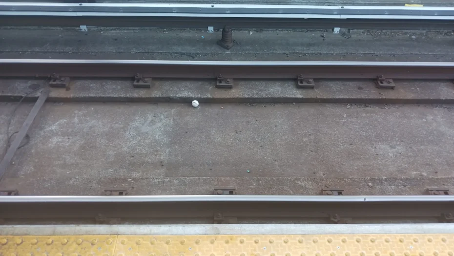 A tennis ball lies on a railway track next to a yellow tactile paving strip.