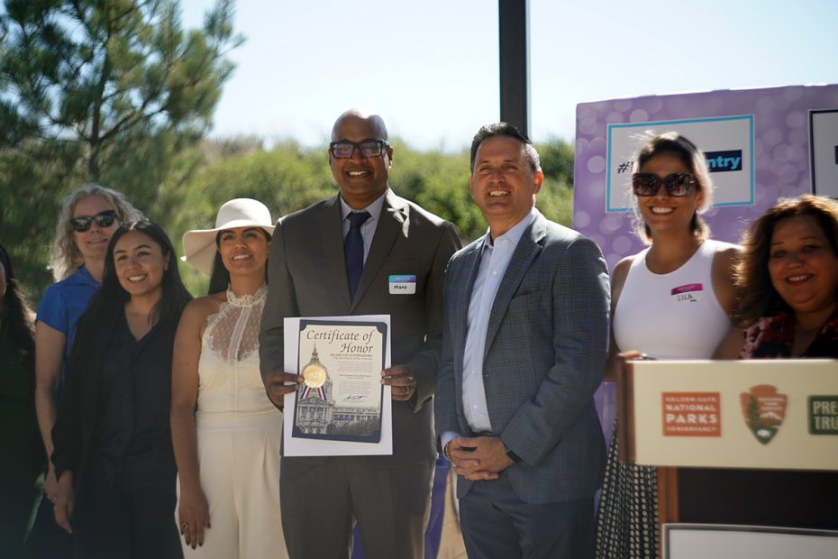 A group of people stands outdoors holding a "Certificate of Honor." They are smiling, dressed in business and casual attire, with trees and a banner in the background.