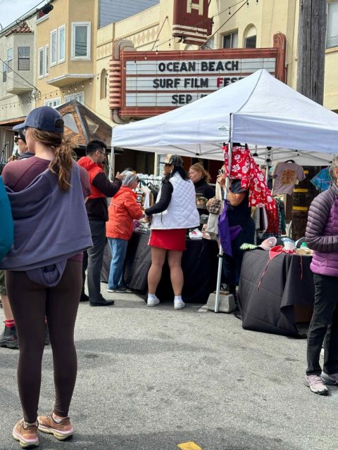 People browsing outdoor market stalls under a canopy near a marquee that reads "Ocean Beach Surf Film Fest Sep." enjoy live performances by Forrest Liu, as the event proudly supports the Stop Asian Hate campaign.