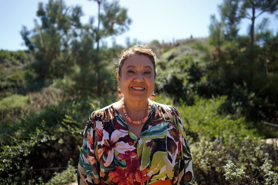 A woman stands outdoors smiling, wearing floral attire and jewelry. She is surrounded by lush greenery and trees under a clear sky.
