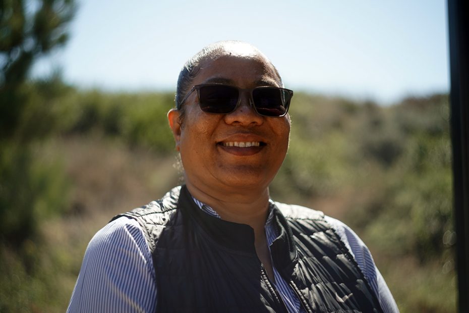 A person wearing sunglasses and a black vest is smiling outdoors with a blurred green landscape in the background.