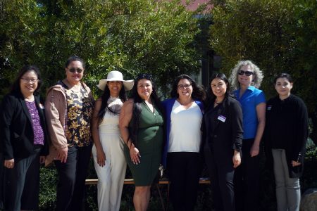 Eight people stand outdoors in front of greenery, some smiling, with a mix of business and casual attire, including sunglasses and a sunhat.