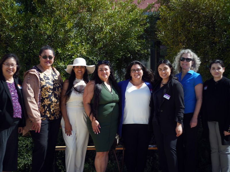 Eight people stand outdoors in front of greenery, some smiling, with a mix of business and casual attire, including sunglasses and a sunhat.