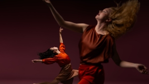 Two people in motion, wearing red and brown outfits, dance against a dark background.