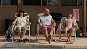 Three people sit on lounge chairs by a pool. The person in the center is looking at the person to their right. It is sunny and there are towels and items around them.