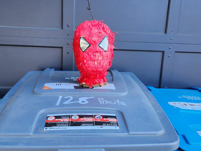 A red piñata shaped like a head with eyes is hanging above a gray recycling bin.