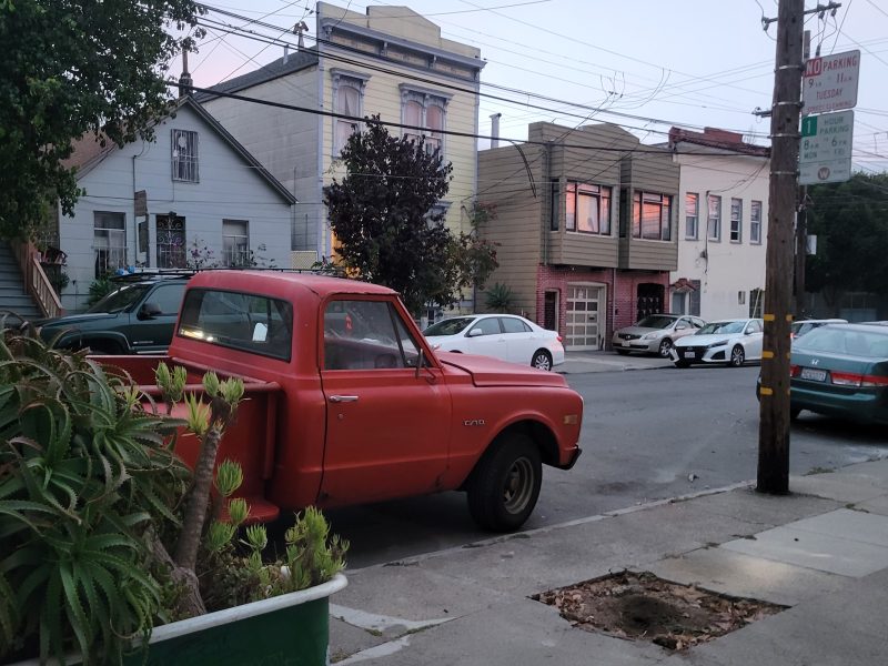 A red vintage pickup truck is parked on a residential street lined with houses. Other cars are parked along the street, and power lines are visible overhead.